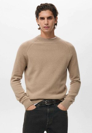 Suéter de punto beige con cuello redondo y puños de canalé, usado sobre jeans oscuros. La textura parece suave con un diseño ajustado clásico.