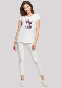 Weißes Baumwoll-T-Shirt mit einem Druck eines Skeletts, das einen rosafarbenen Herzballon hält, kombiniert mit weißen Skinny-Jeans und weißen Sneakers.