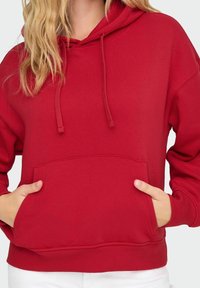 Femme portant un hoodie rouge avec un cordon de serrage, les mains dans la poche avant, et un pantalon blanc visible en bas.