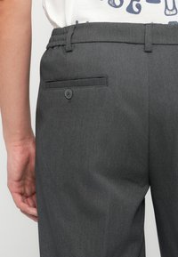 Πίσω όψη ατόμου που φοράει σκούρα γκρι ραμμένο παντελόνι με πίσω τσέπη με κουμπί και ελαστική μέση, το χέρι ακουμπάει δίπλα στο ισχίο.