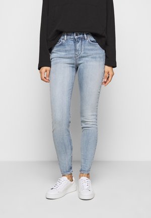 Person iført lyseblå skinny jeans, hvide sneakers og en sort langærmet top mod en ensfarvet grå baggrund.