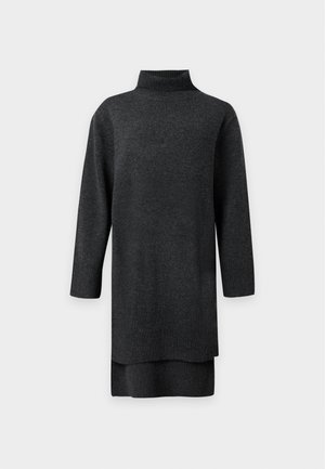 Un mannequin porte une robe-pull oversize à col roulé, gris anthracite, avec un ourlet asymétrique plus court devant et plus long derrière, ainsi que des bottines noires. Il se tient devant un fond clair uni.