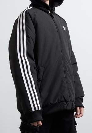 Personne portant une veste noire Adidas avec trois bandes blanches sur les manches et une capuche, debout devant un fond clair uni.