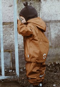Bruine waterdichte jas en bijpassende broek met elastische boorden. Kind draagt een donkerbruine hoed met kleine oortje-achtige versieringen.