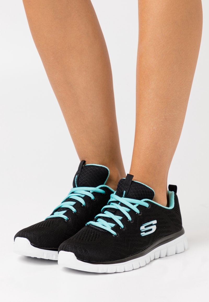 Pieds chaussés de baskets noires à semelles blanches et lacets bleu clair, posés sur une surface blanche.
