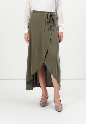 Mujer con una falda envolvente verde oliva con un lazo en la cintura, blusa de mangas largas transparente y estampada, y tacones de punta taupe.