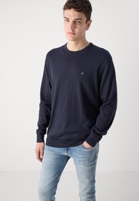 Tamsiai mėlynas medvilninis megztinis ilgomis rankovėmis, su raukiniais rankogaliuose ir maža išsiuvinėta logo ant krūtinės; derinama su šviesiai mėlynomis džinsomis.