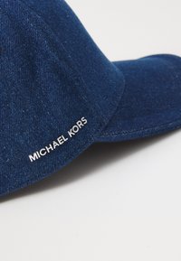 Modrá denimová kšiltovka s zakřiveným kšiltem a vyšívaným bílým logem s nápisem "MICHAEL KORS" na boku. Hladká textura, neformální design.