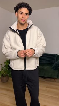 Φλις μπουφάν σε κρεμ χρώμα με φερμουάρ μπροστά, ψηλή γιακά και μαύρο περίγραμμα. Συνδυασμένο με μαύρη φόρμα. Εσωτερικός χώρος με πράσινο καναπέ και φυτά.