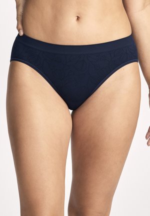 Frau trägt marineblaue Unterwäsche mit Blumenmuster, zeigt den Bauchbereich und die oberen Oberschenkel vor einem schlichten Hintergrund.