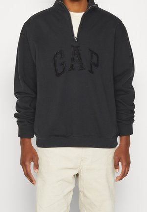 Hombre con sudadera negra GAP de media cremallera con gran logo en relieve, combinada con pantalones beige claro, posando frente a un fondo liso.