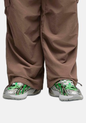 Le sneakers presentano un design verde e argento con motivi fluidi, abbinate a pantaloni marroni larghi, che mostrano uno stile rilassato.