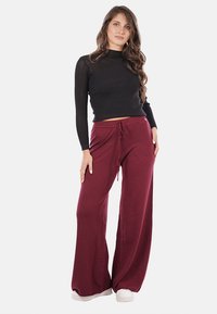 Top lungo a maniche lunghe nero a coste abbinato a pantaloni larghi bordeaux con vita regolabile. Il modello si trova su uno sfondo uniforme.