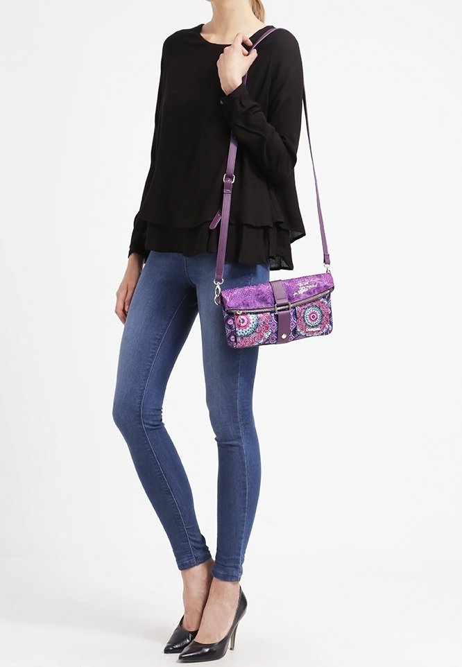 Lilla crossbody taske med unikke cirkulære mønstre, metalliske detaljer og snap-lukning. Båret sammen med en sort lagdelt top og blå skinny jeans.