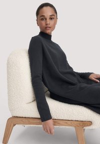 Schwarzer Rollkragenpullover aus weichem Material, mit langen Ärmeln, lockerer Passform und hohem Kragen, kombiniert mit einem strukturierten cremefarbenen Sessel.