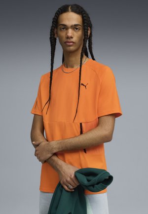 Nuori henkilö, jolla on pitkät letit, wear oranssia Puma-paitaa ja pitää vihreää hupparia vaaleanharmaata taustaa vasten.