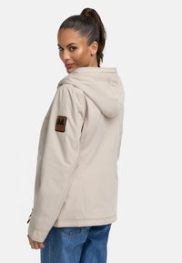 Beige hooded jacket with a textured fabric and a brown logo patch on the left shoulder. Features a relaxed fit and visible stitching details.