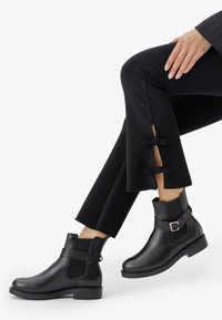 Mujer con pantalones negros cortos con lazadas laterales y botas de cuero negro hasta el tobillo con hebillas, sentada con las piernas cruzadas.