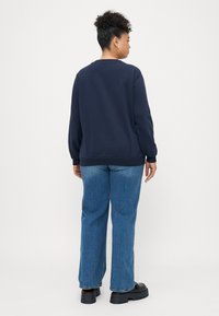 Námornícky modrý sveter s rebrovanými manžetami a spodným lemom, v kombinácii s rovnými svetlomodrými džínami. Model zobrazený zozadu.