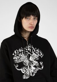 Black zip-up hoodie featuring a white graphic print of an eagle design on the chest, with a drawstring hood and soft fabric texture.