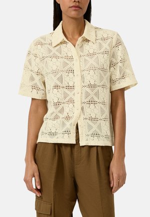 Vrouw draagt een crème-kleurige, korte mouw, gehaakte blouse met knopen, gecombineerd met bruine, high-waisted cargobroek, staand tegen een effen achtergrond.