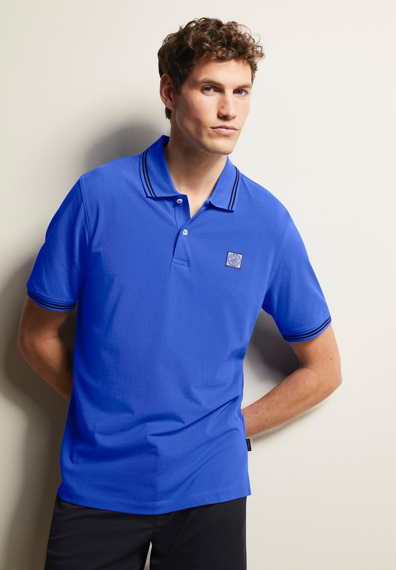 Blaues Polohemd aus strukturiertem Material, mit zwei schwarzen Streifen am Kragen und an den Ärmeln sowie einem kleinen Logo auf der Brust. Kurzarm-Design.