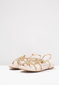 Ploché sandály s růžovými podrážkami a zlatými, klenotem zdobenými květinovými pásky, se zapínáním na kotníku, vystavené na bílé ploše s jednobarevným pozadím.