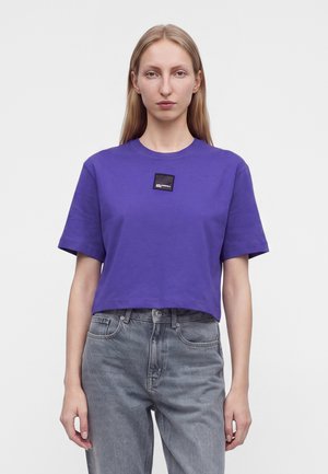 Kvinde iført en lilla cropped t-shirt med en lille sort lap på brystet og falmede grå jeans, stående mod en ensfarvet baggrund.