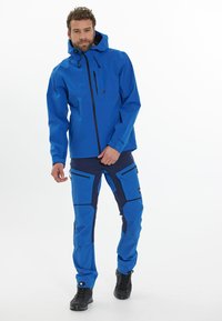 Sininen vettä hylkivä takki hupulla ja vetoketjulla, yhdistettynä sinisiin ja tummansinisiin housuihin, joissa on viistoja taskuja ja kestävä rakenne.