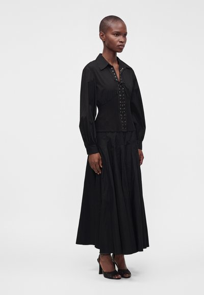 Femme portant une longue robe noire avec un haut style corset à lacets et des manches longues bouffantes, debout en talons noirs ouverts à l'avant sur un fond uni.
