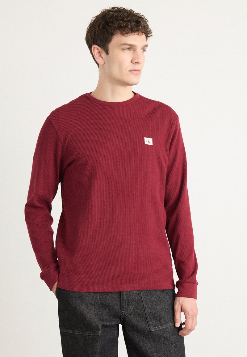 Bordeaux langemouws heren overhemd in ribbeltextuur met een ronde hals en een logo-opdruk op de linkerkant van de borst. Gecombineerd met donkerdenim broek.
