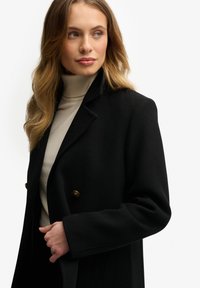 Zwarte wollen jas met een op maat gemaakt ontwerp, dubbel gekregen voorkant en goudkleurige knopen, gelaagd over een lichtbeige coltrui.