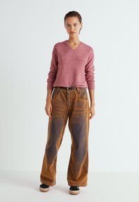 Rosa V-Ausschnitt-Strickpullover mit geripptem Muster; weite brauneCordhosen mit Knopfleiste, Seitentaschen und vertikalen Musterelementen.