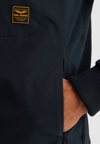 Felpa con cappuccio blu scuro in tessuto testurizzato, cuciture visibili, un'etichetta logata sul petto e un dettaglio con tasca laterale con zip.