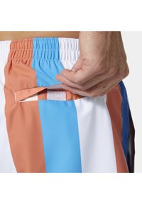 Ręka otwierająca tylną kieszeń na rzep w wielokolorowych szortach w niebieskie, białe i pomarańczowe paski z elastycznym pasem.