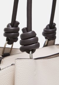 Sac à main en cuir beige clair avec finition texturée, doté de poignées nouées marron foncé et d'accents en métal. Ouverture zippée sur le dessus.