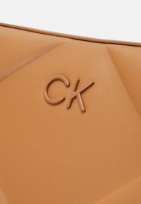 Beżowa torba z ekoskóry o pikowanej fakturze i wytłoczonym logo CK. Posiada ukośne przeszycia i gładką powierzchnię.