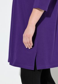 Lila tunika-klänning i mjukt tyg, med korta ärmar, sidofrakar vid fållen och A-linjeform. Bärs över svarta leggings.