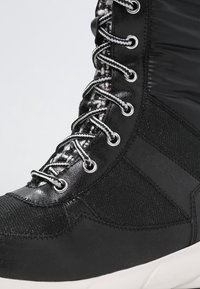 Baskets montantes noires en tissu texturé, avec des superpositions brillantes, des lacets rayés blancs et noirs, et une semelle en caoutchouc blanche.