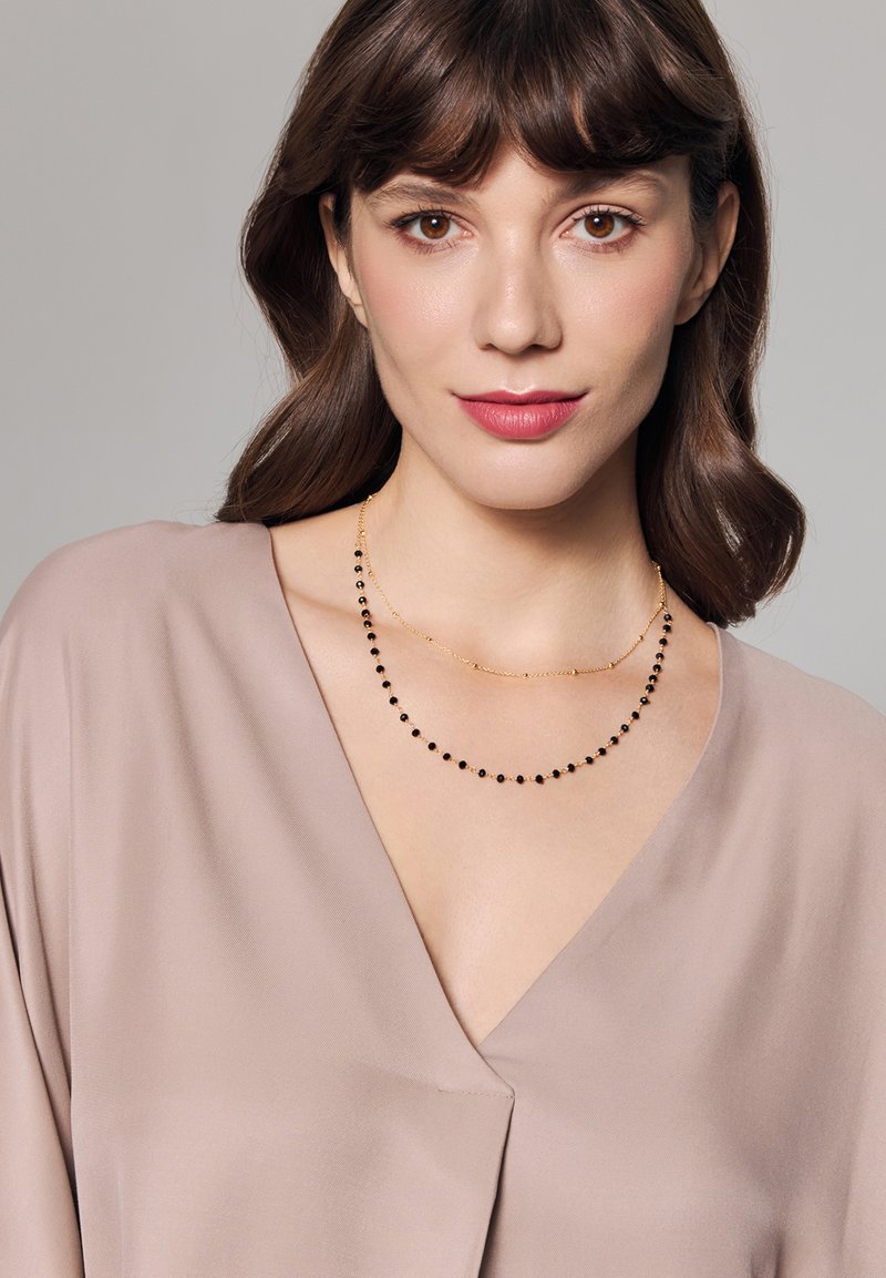 Złoty naszyjnik z dwiema warstwami, z jedną składającą się z małych czarnych koralików, a drugą z wydłużonych ogniw, noszony na beżowej bluzce.