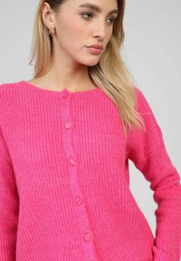 Cardigan en tricot rose avec un col rond, des manches longues et cinq boutons assortis sur le devant. Tissu texturé avec une apparence douce.