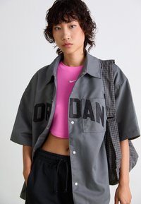 Gray short-sleeve shirt with black "OR DAN" lettering, worn over a pink fitted top by Nike; paired with black sweatpants and a checkered bag.