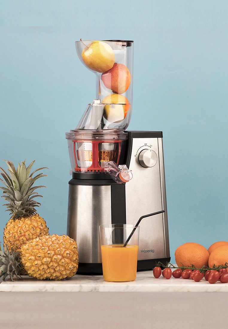 Entsafter mit Edelstahl- und transparenten Kunststoffkomponenten. Äpfel im Trichter, Saft fließt in ein Glas mit einem schwarzen Strohhalm. Ananas und Früchte daneben.