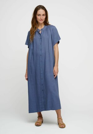 Femme portant une robe bleue ample à manches courtes avec des boutons sur le devant et des sandales à bride à la cheville, debout devant un fond uni.
