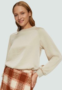 Hellbeige Sweatshirt mit Rundhalsausschnitt und Raglanärmeln. Weicher Stoff mit gerafften Bündchen. Kombiniert mit einem gemusterten Karottenrock in warmen Tönen.