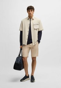 Camicia beige a maniche corte con taschino, camicia blu scuro a maniche lunghe sotto, pantaloni corti beige, porta una borsa a tracolla nera e blu e indossa scarpe da ginnastica nere.
