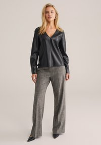 Schwarze Leder-Bluse mit langen Ärmeln und V-Ausschnitt, kombiniert mit grauen, karominierten, weiten Hosen und spitzen schwarzen High Heels. Glatte Textur und taillierter Schnitt.