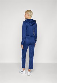 Zils sporta treniņtērps ar kapuci, ar garām piedurknēm un pieguļošām biksēm. Audums izskatās gluds, un tas ir kopā ar baltām sporta kurpēm.