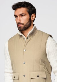 Hombre con cabello oscuro y barba, vistiendo un chaleco acolchado beige sobre un suéter blanco de punto con cable, mirando ligeramente hacia la izquierda sobre un fondo liso.