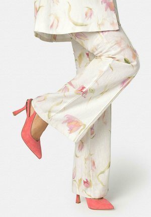 Pantaloni bianchi a fiori abbinati a décolleté slingback in camoscio rosa corallo, un piede sollevato dietro l'altro in posa a metà passo.